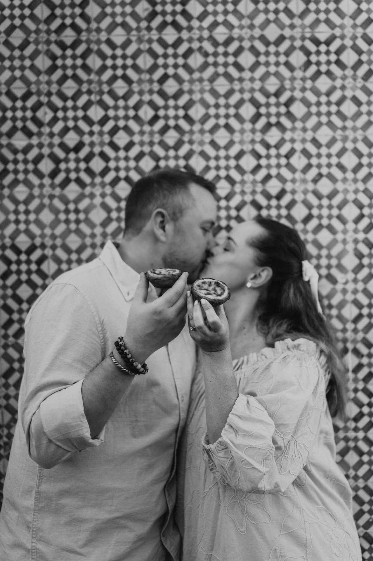 Black-and-white photograph: the couple leaning together with custard tarts held close to their faces against the patterned tile wall during their engagement photoshoot in Lisbon.