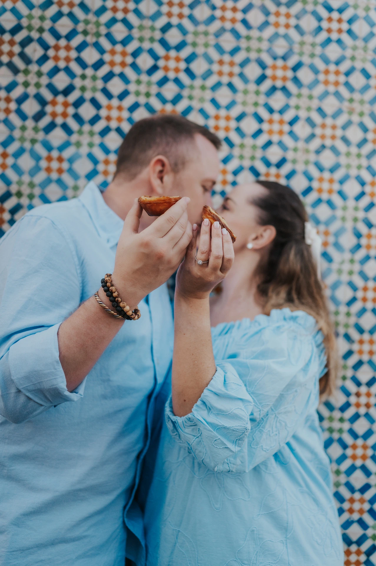 The couple leaning together with custard tarts held close to their faces against the patterned tile wall during their engagement photoshoot in Lisbon.