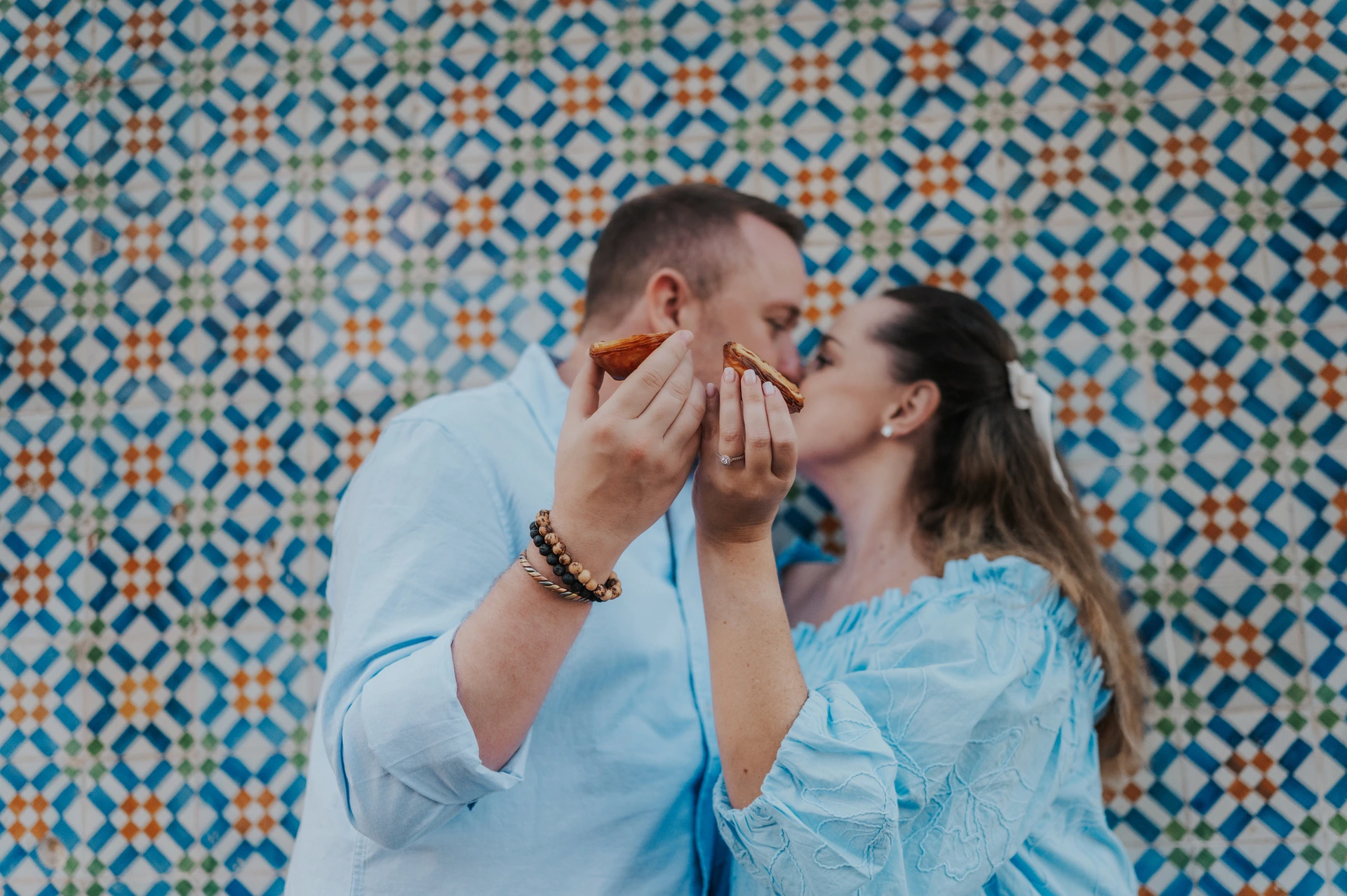 The couple leaning together with custard tarts held close to their faces against the patterned tile wall during their engagement photoshoot in Lisbon.