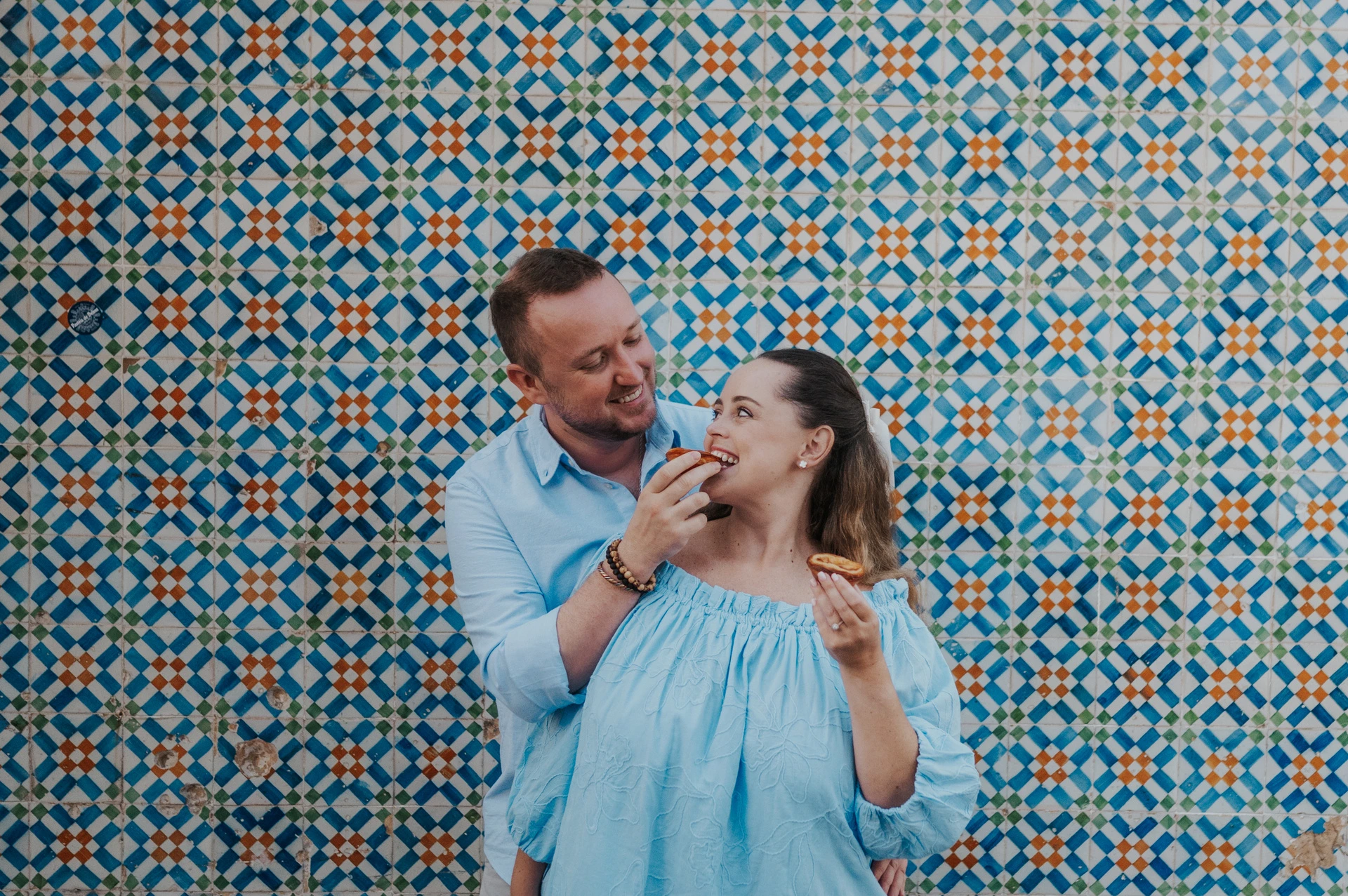 The man holding a custard tart towards his smiling partner beside the tile wall during their engagement photoshoot in Lisbon.