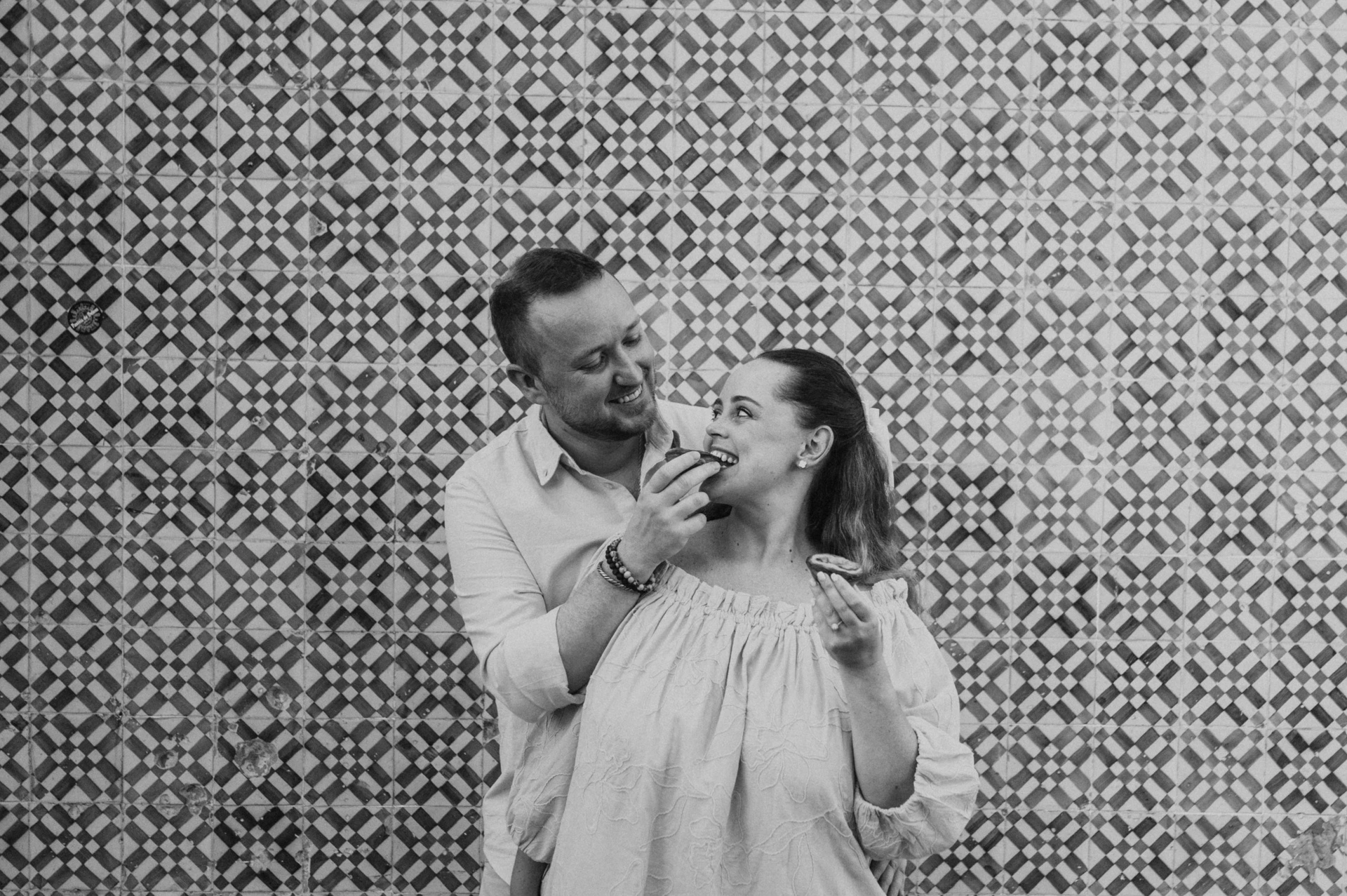 Black-and-white photograph: the man holding a custard tart towards his smiling partner beside the tile wall during their engagement photoshoot in Lisbon.