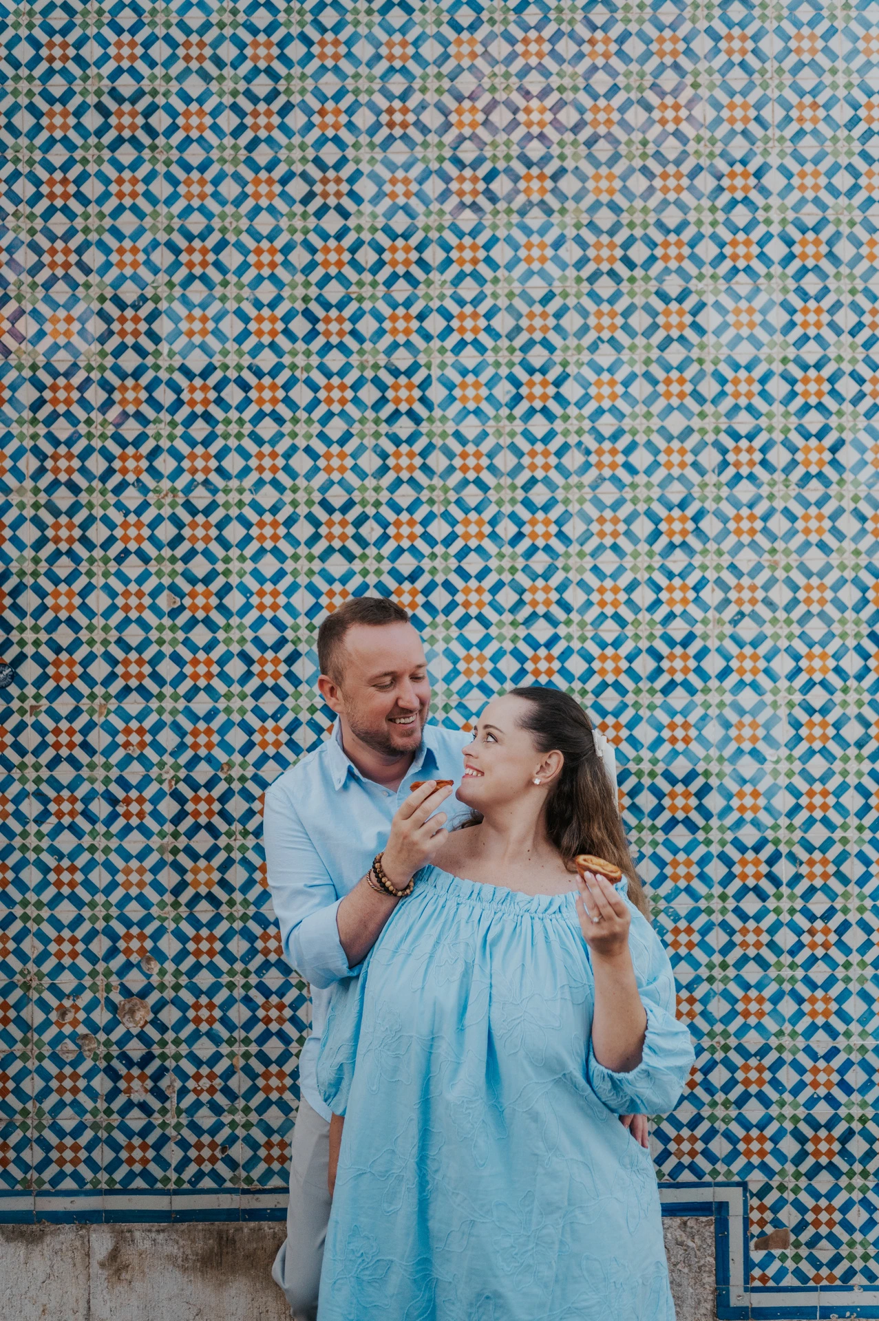 The man holding a custard tart towards his smiling partner beside the tile wall during their engagement photoshoot in Lisbon.
