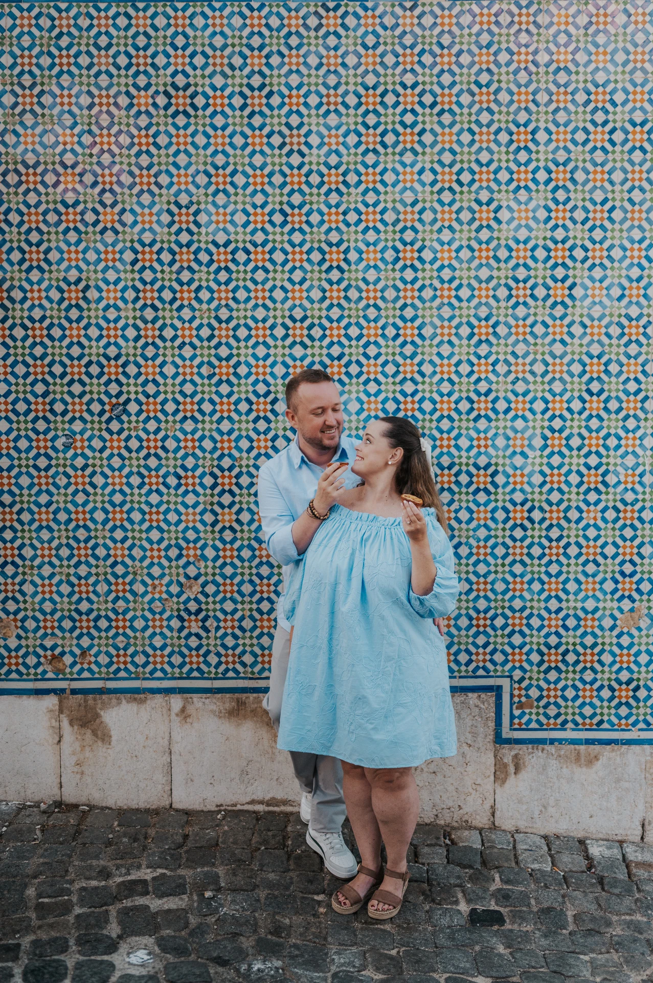 The man holding a custard tart towards his smiling partner beside the tile wall during their engagement photoshoot in Lisbon.