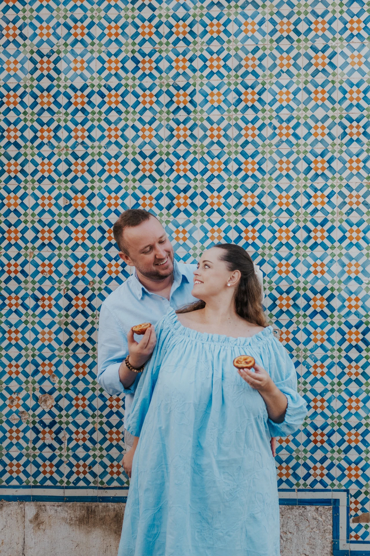 The man holding a custard tart towards his smiling partner beside the tile wall during their engagement photoshoot in Lisbon.