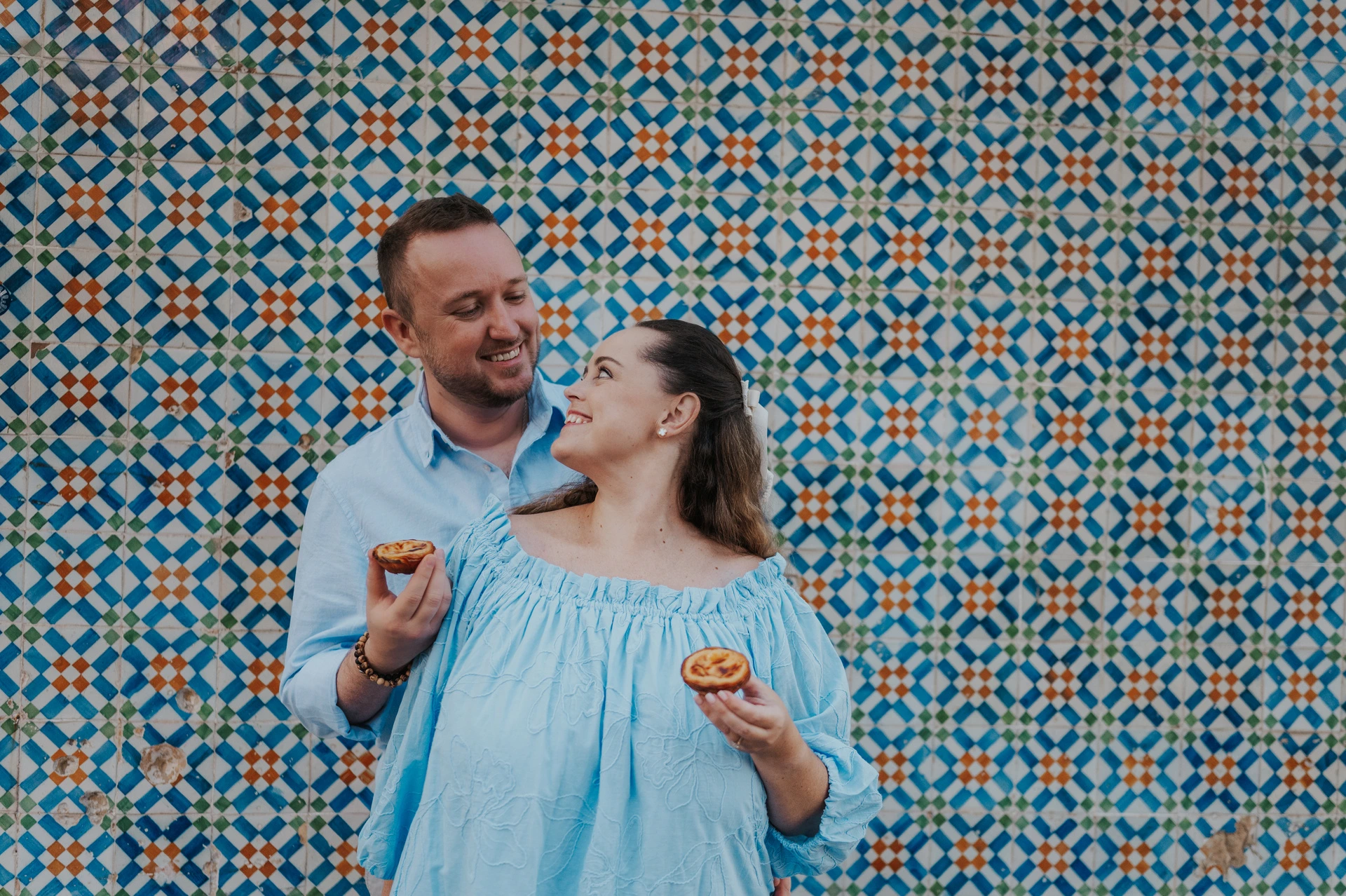 The man holding a custard tart towards his smiling partner beside the tile wall during their engagement photoshoot in Lisbon.