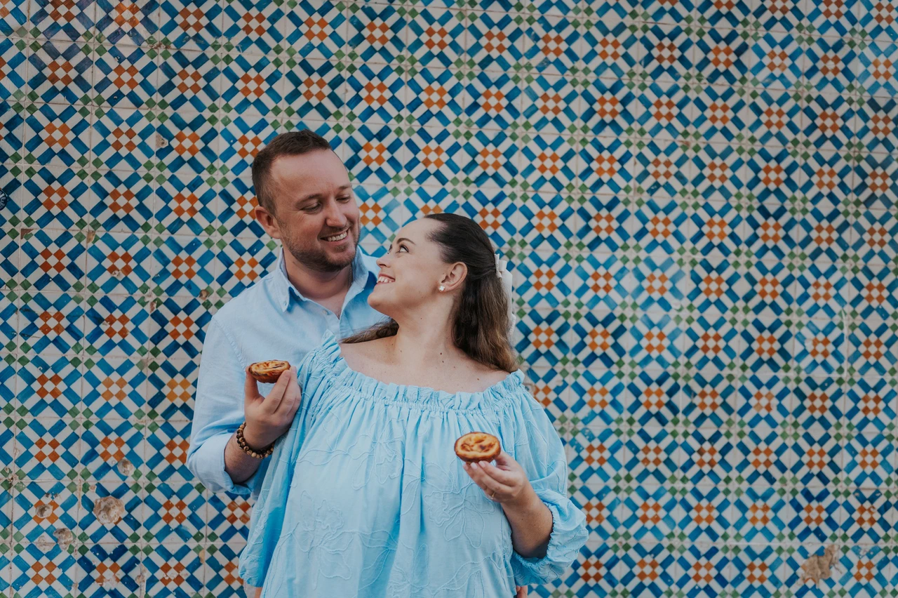 Sharing a pastry and a smile | Lisbon engagement photoshoot The man holding a custard tart towards his smiling partner beside the tile wall during their engagement photoshoot in Lisbon.