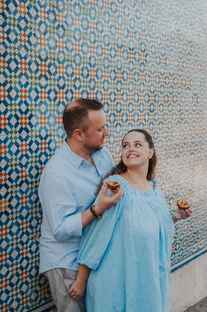 Sharing a pastry and a smile | Lisbon engagement photoshoot The man holding a custard tart towards his smiling partner beside the tile wall during their engagement photoshoot in Lisbon.