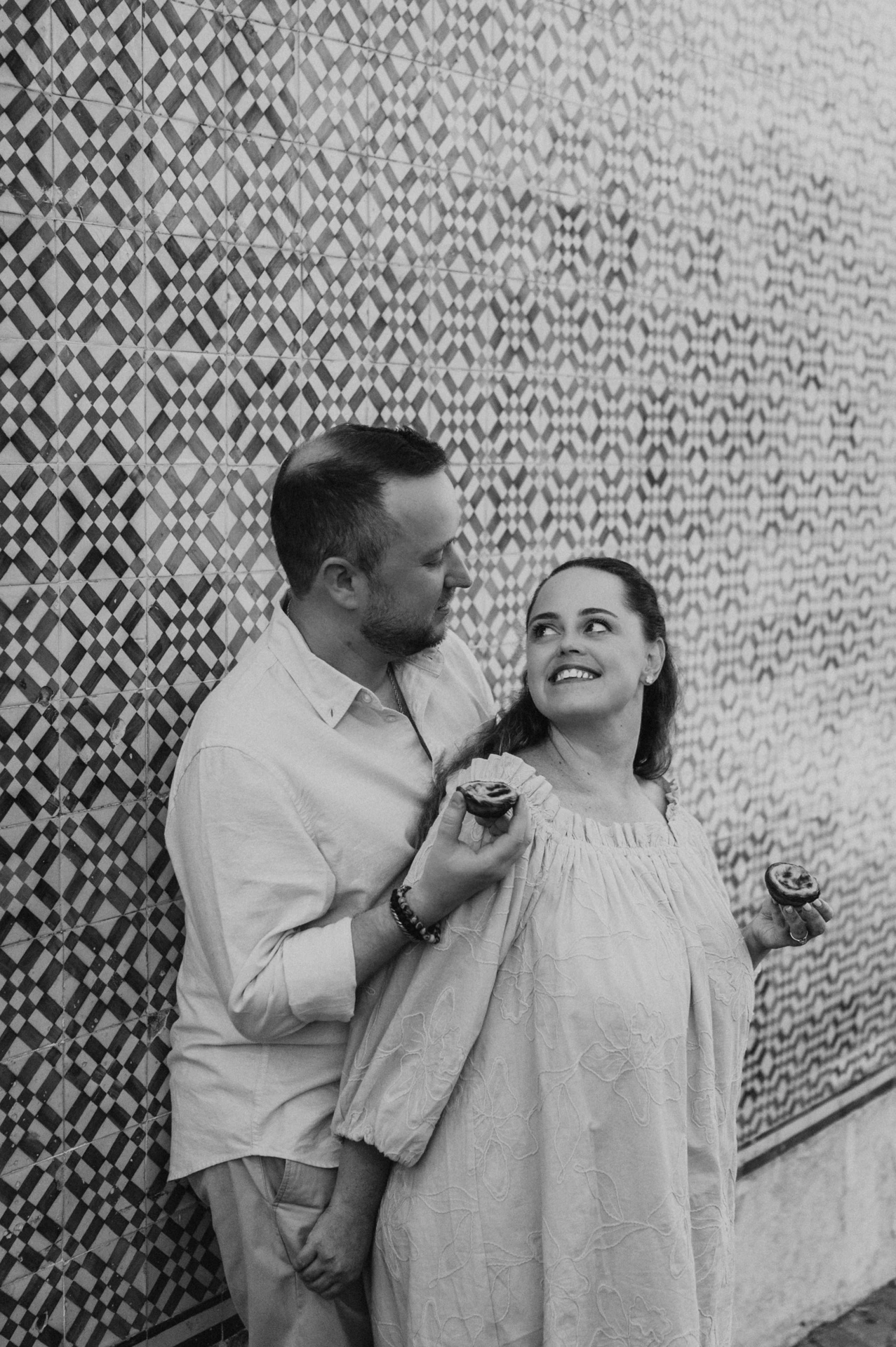 Black-and-white photograph: the man holding a custard tart towards his smiling partner beside the tile wall during their engagement photoshoot in Lisbon.