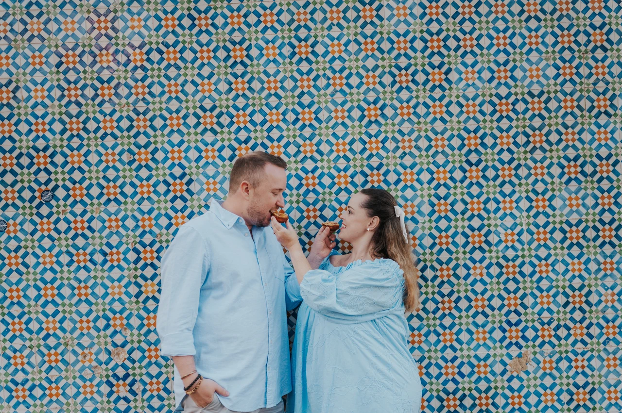 Offering each other a bite of pastéis | Lisbon engagement photoshoot The woman holding a custard tart towards her partner in front of the patterned tiles during their engagement photoshoot in Lisbon.