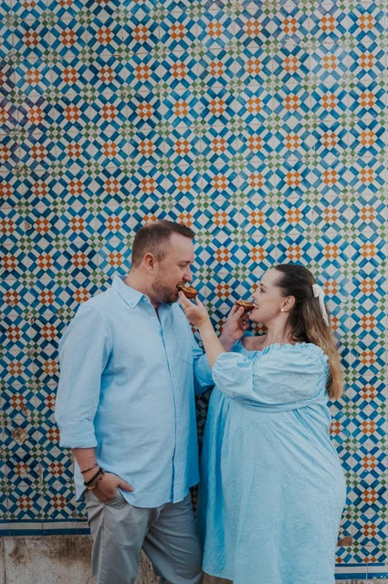 Offering each other a bite of pastéis | Lisbon engagement photoshoot The woman holding a custard tart towards her partner in front of the patterned tiles during their engagement photoshoot in Lisbon.