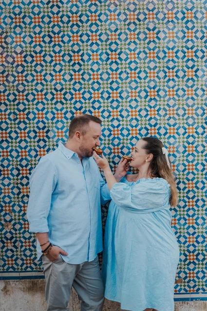 Offering each other a bite of pastéis | Lisbon engagement photoshoot The woman holding a custard tart towards her partner in front of the patterned tiles during their engagement photoshoot in Lisbon.