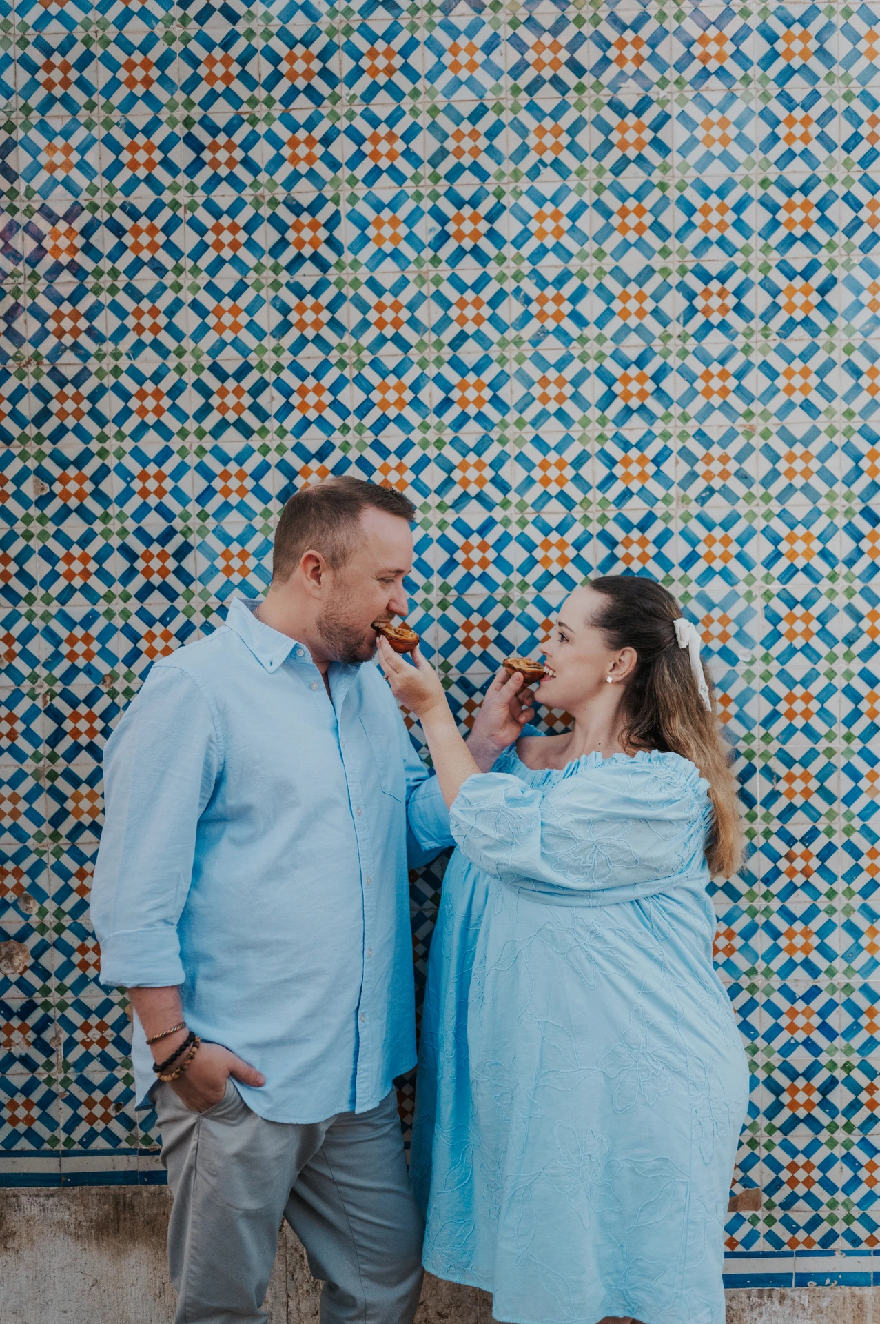 The woman holding a custard tart towards her partner in front of the patterned tiles during their engagement photoshoot in Lisbon.