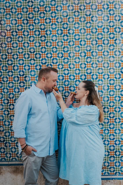Offering each other a bite of pastéis | Lisbon engagement photoshoot The woman holding a custard tart towards her partner in front of the patterned tiles during their engagement photoshoot in Lisbon.
