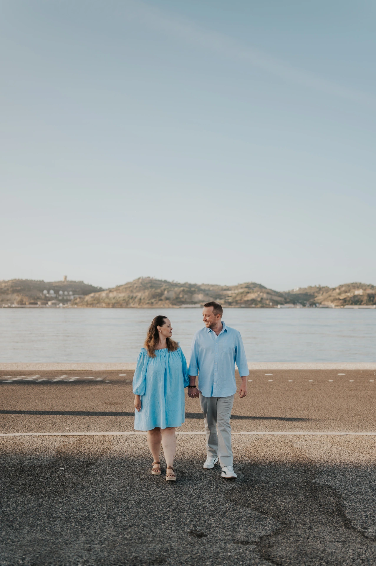 The couple walking hand in hand with the river stretching out behind them during their engagement photoshoot in Lisbon.