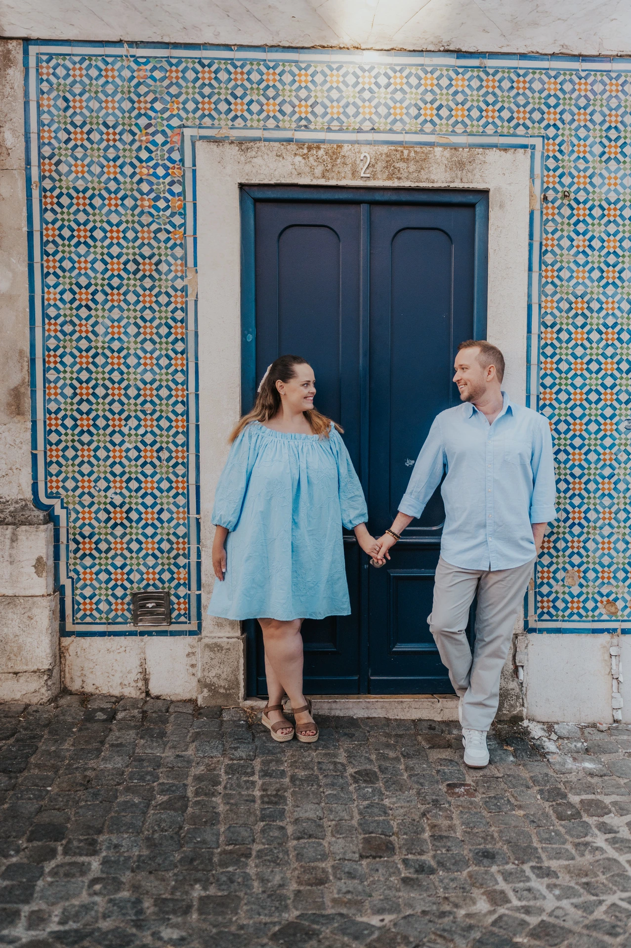 The couple holding hands on either side of a blue doorway surrounded by patterned tiles during their engagement photoshoot in Lisbon.
