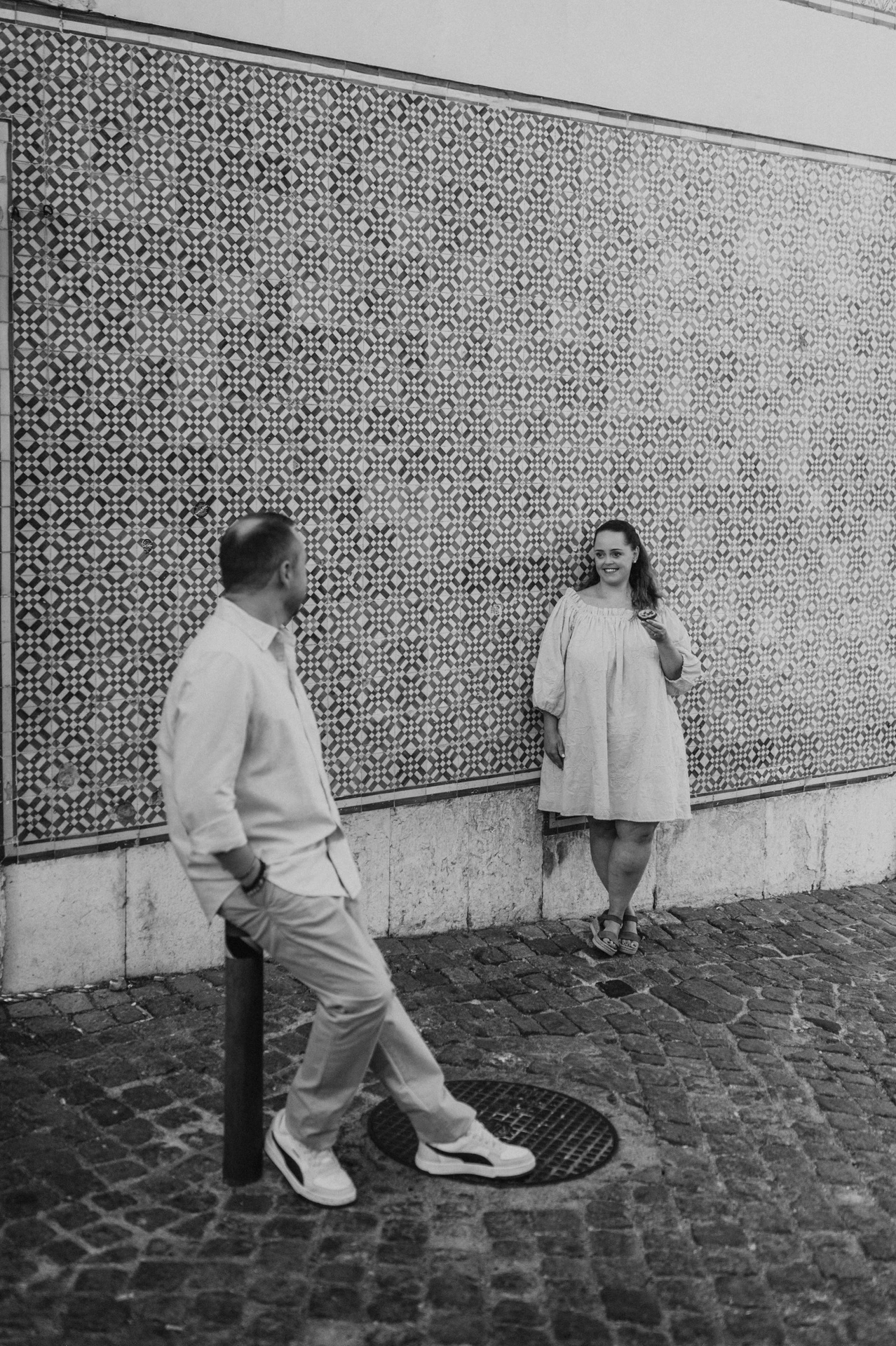 Black-and-white photograph: the couple leaning apart against the long patterned tile wall during their engagement photoshoot in Lisbon.