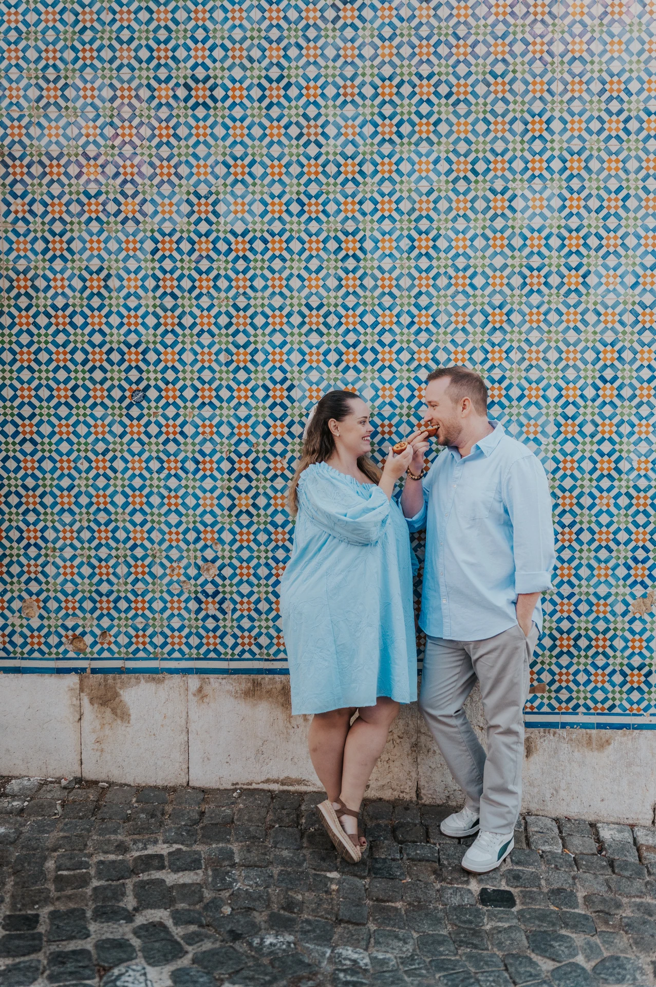 The couple standing close together and smiling with pastries by the tile wall during their engagement photoshoot in Lisbon.