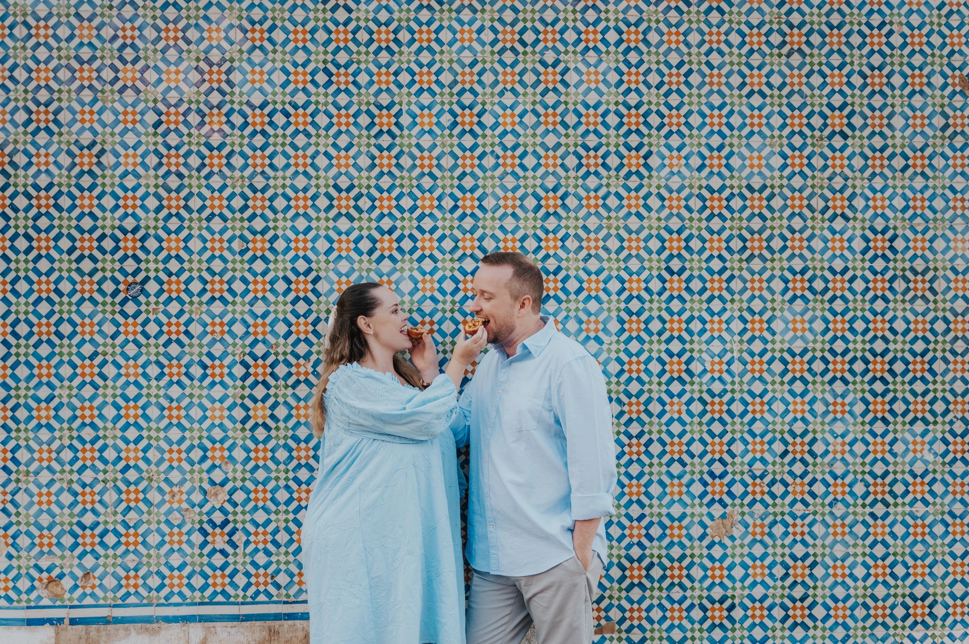 The couple standing close together and smiling with pastries by the tile wall during their engagement photoshoot in Lisbon.