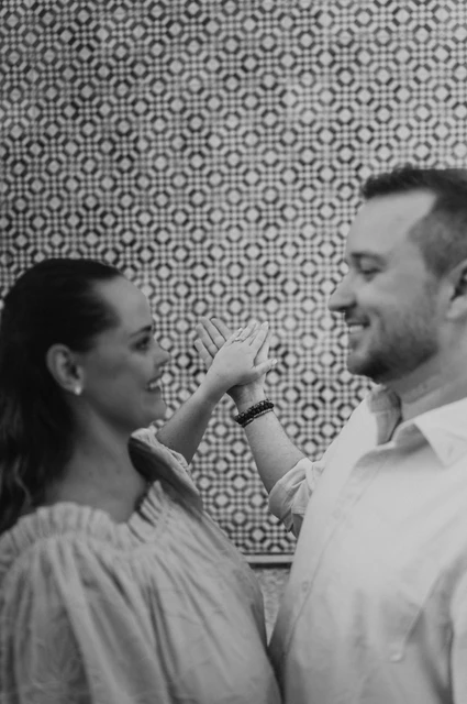 A playful hand-in-hand portrait in black and white | Lisbon engagement photoshoot Black-and-white photograph: the couple facing each other with their joined hands raised near their faces by the tile wall during their engagement photoshoot in Lisbon.