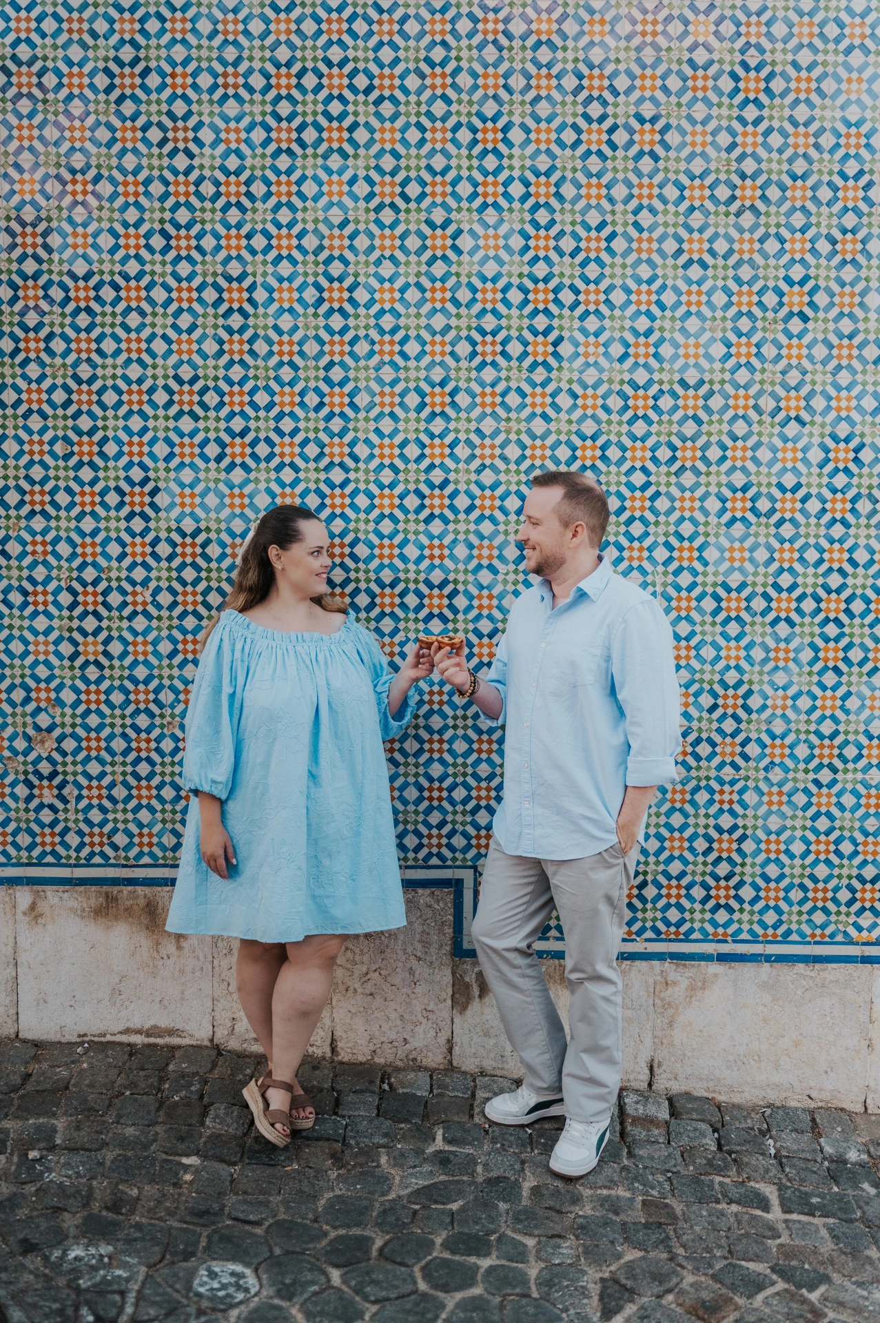 The couple smiling together with pastries in front of a blue and yellow patterned tile wall during their engagement photoshoot in Lisbon.