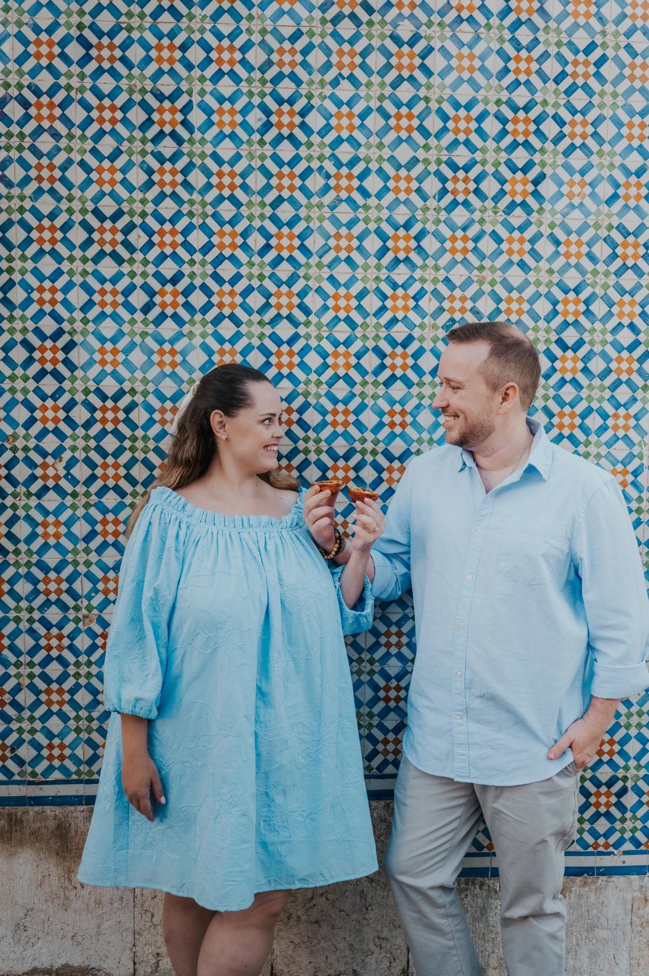 The couple smiling together with pastries in front of a blue and yellow patterned tile wall during their engagement photoshoot in Lisbon.