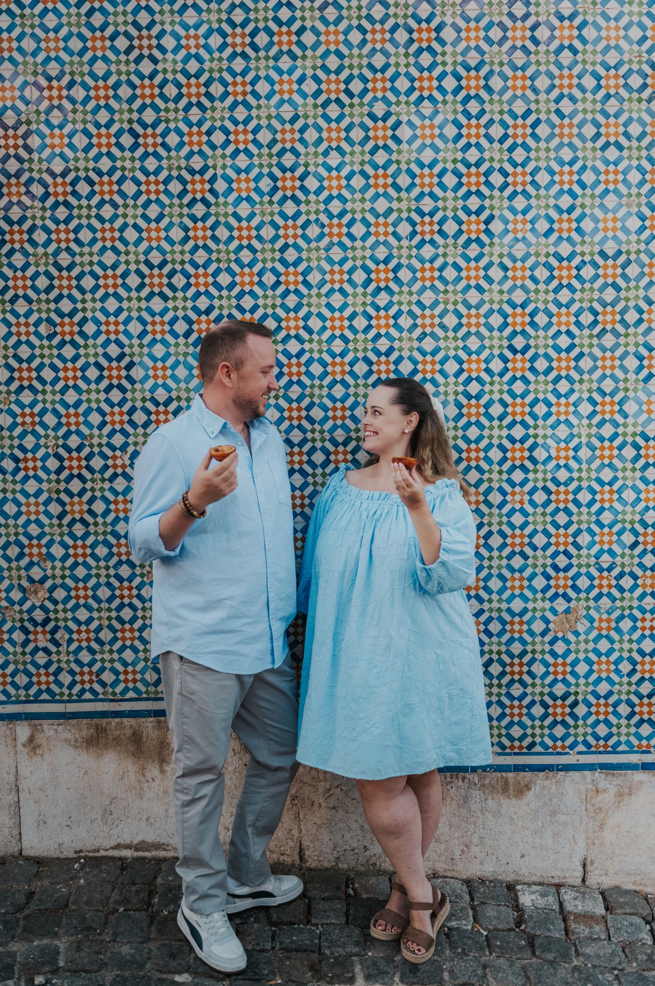 The couple smiling together with pastries in front of a blue and yellow patterned tile wall during their engagement photoshoot in Lisbon.