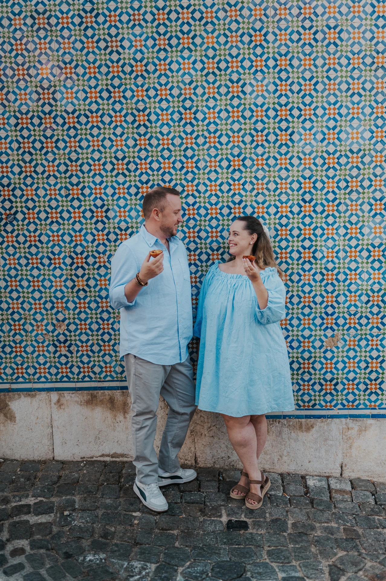 The couple smiling together with pastries in front of a blue and yellow patterned tile wall during their engagement photoshoot in Lisbon.