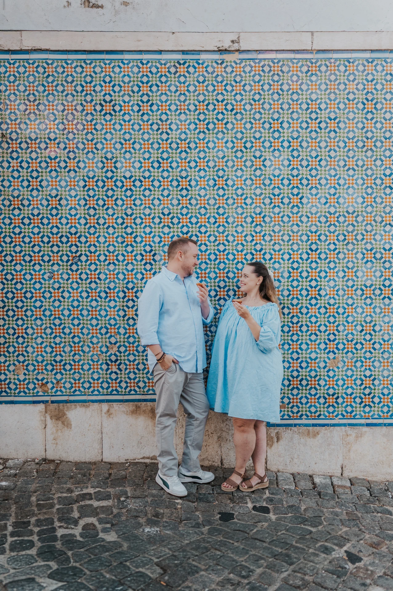 The couple smiling together with pastries in front of a blue and yellow patterned tile wall during their engagement photoshoot in Lisbon.