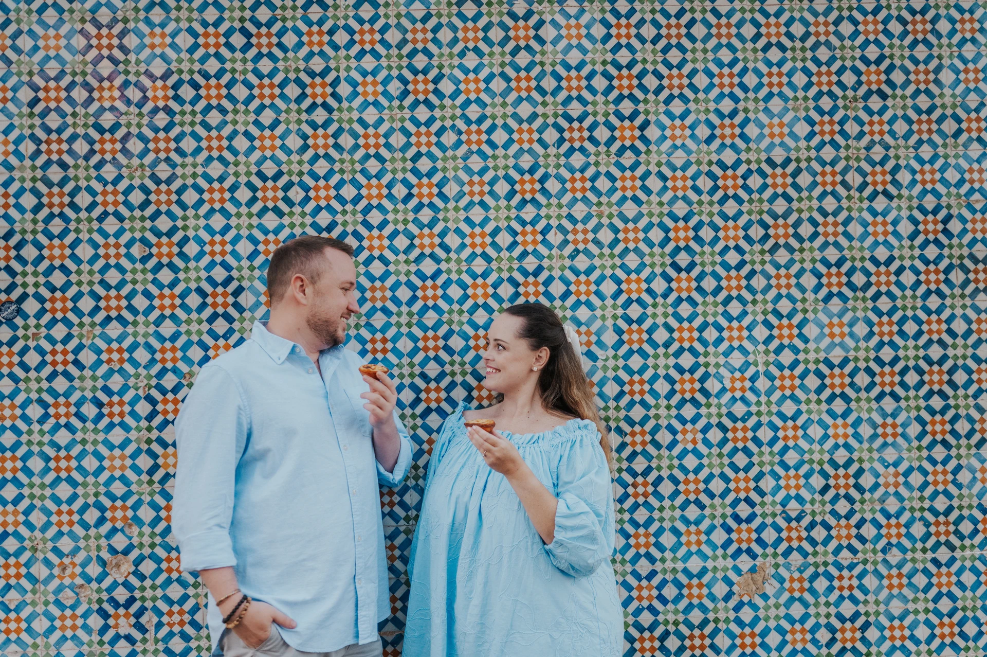 The couple smiling together with pastries in front of a blue and yellow patterned tile wall during their engagement photoshoot in Lisbon.