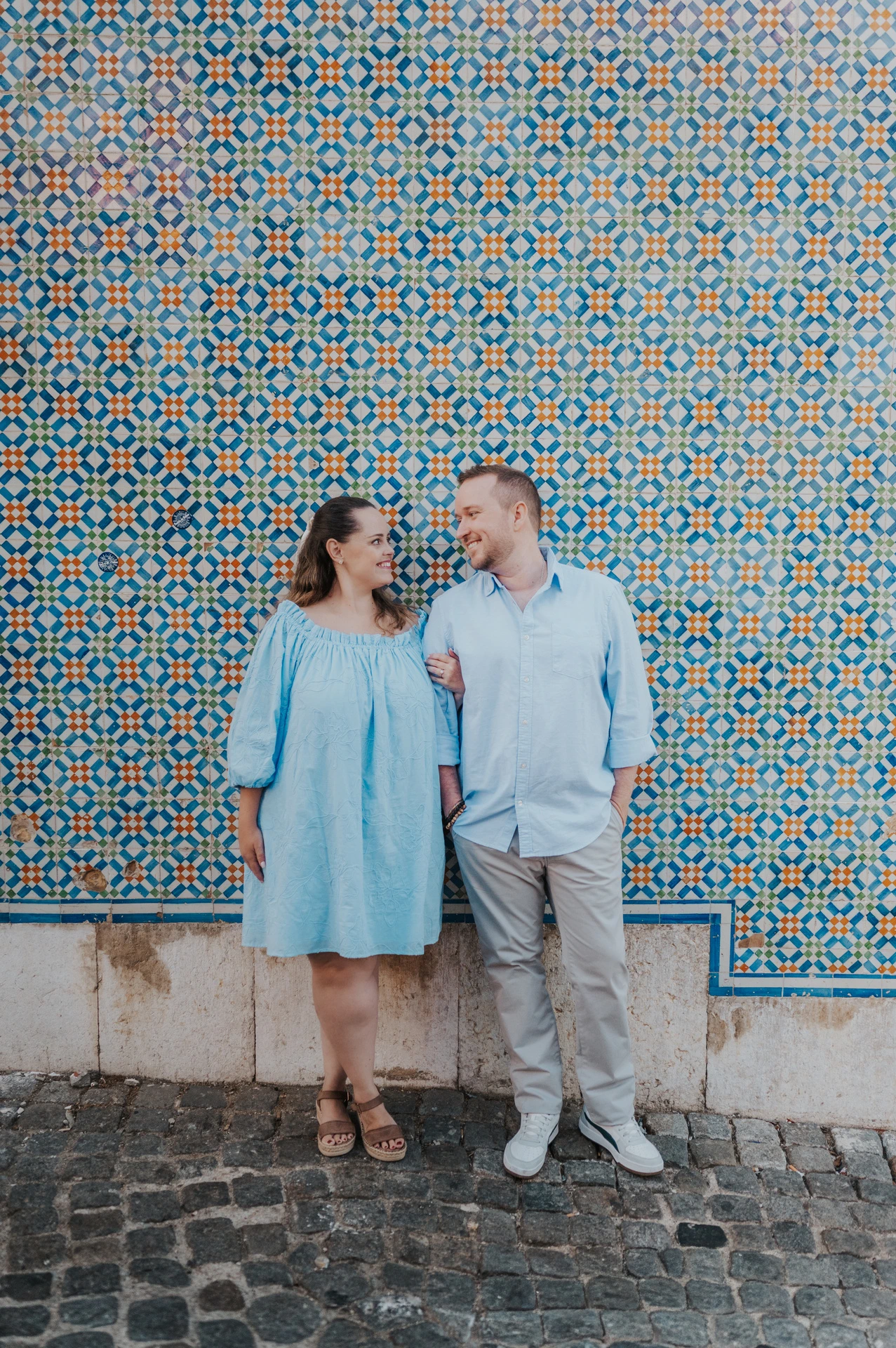 The couple smiling together with pastries in front of a blue and yellow patterned tile wall during their engagement photoshoot in Lisbon.