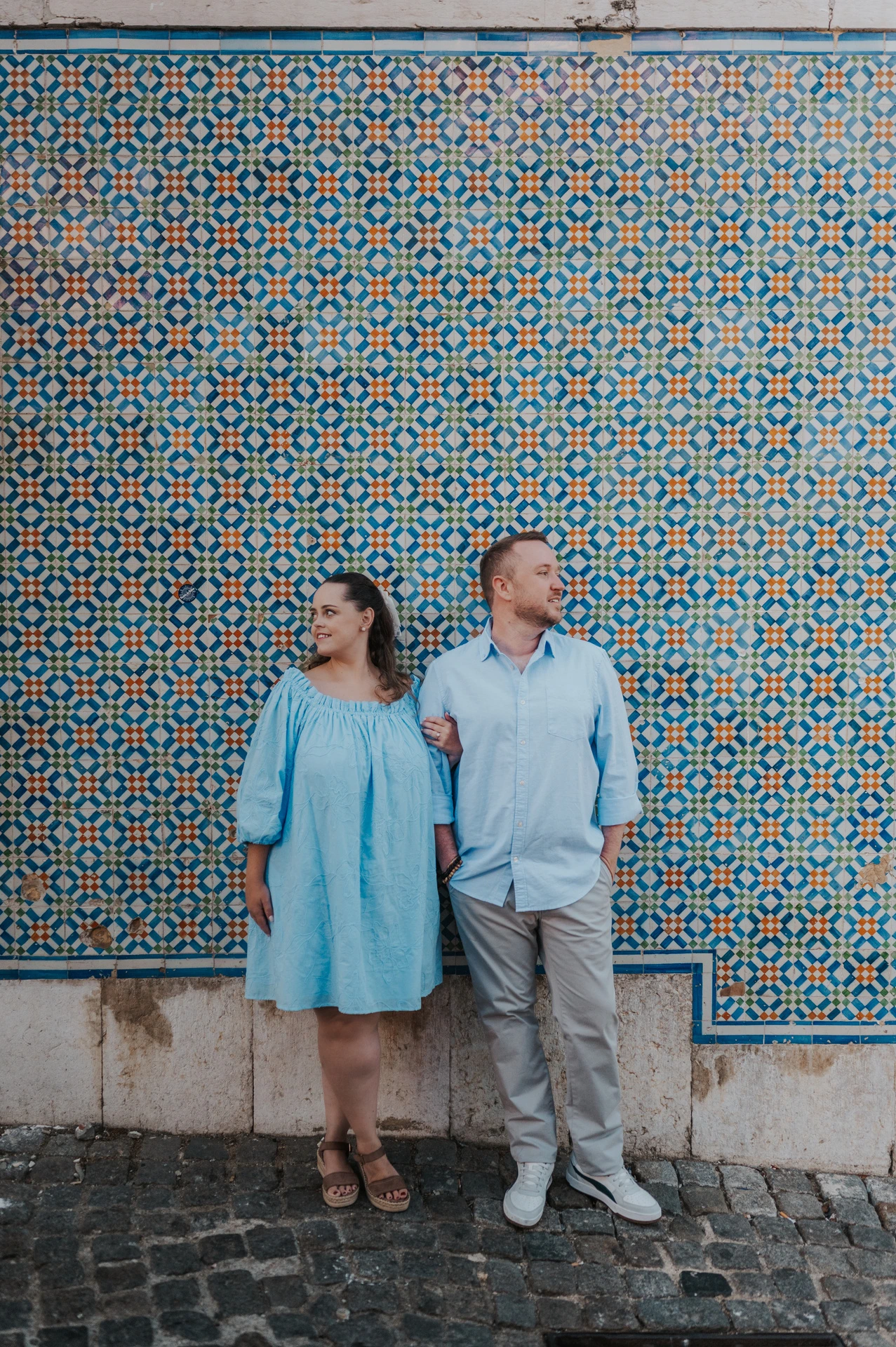 The couple smiling together with pastries in front of a blue and yellow patterned tile wall during their engagement photoshoot in Lisbon.