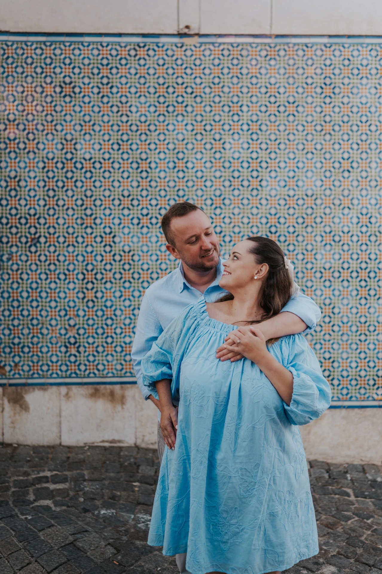 The man with an arm around his partner’s shoulder as they smile at each other against the tiles during their engagement photoshoot in Lisbon.