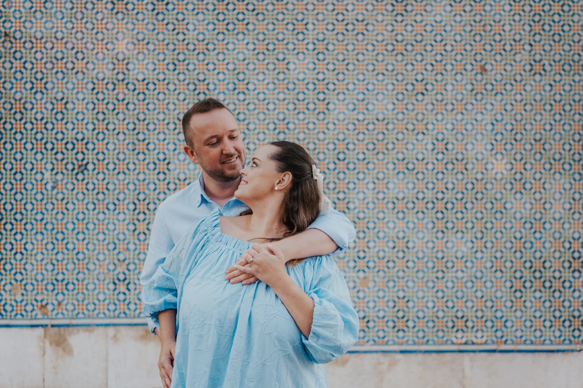 The man with an arm around his partner’s shoulder as they smile at each other against the tiles during their engagement photoshoot in Lisbon.