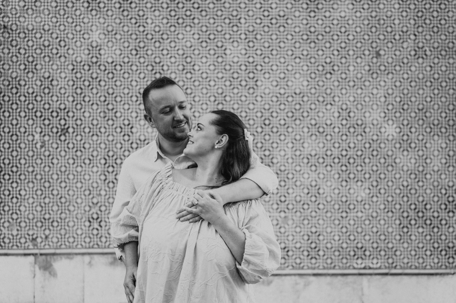 Black-and-white photograph: the man with an arm around his partner’s shoulder as they smile at each other against the tiles during their engagement photoshoot in Lisbon.