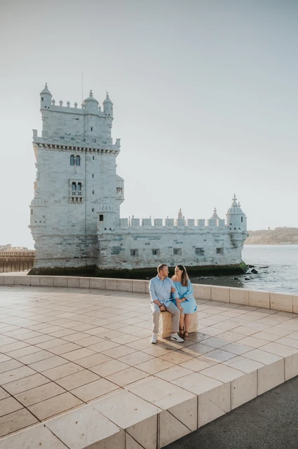 A seated portrait beside Belém Tower | Lisbon engagement photoshoot The woman seated on a stone block with her partner close behind her and Belém Tower in the background during their engagement photoshoot in Lisbon.
