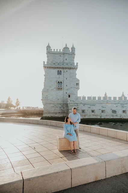 The woman seated on a stone block with her partner close behind her and Belém Tower in the background during their engagement photoshoot in Lisbon.