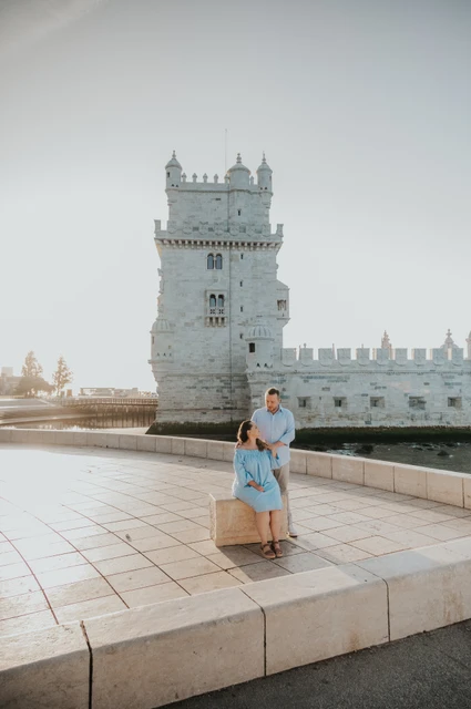 A seated portrait beside Belém Tower | Lisbon engagement photoshoot The woman seated on a stone block with her partner close behind her and Belém Tower in the background during their engagement photoshoot in Lisbon.