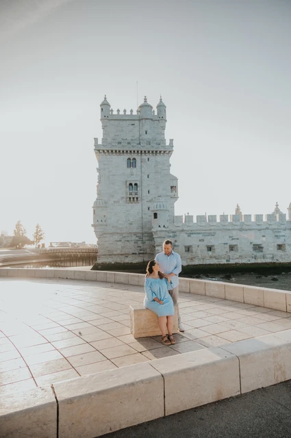 A seated portrait beside Belém Tower | Lisbon engagement photoshoot The woman seated on a stone block with her partner close behind her and Belém Tower in the background during their engagement photoshoot in Lisbon.