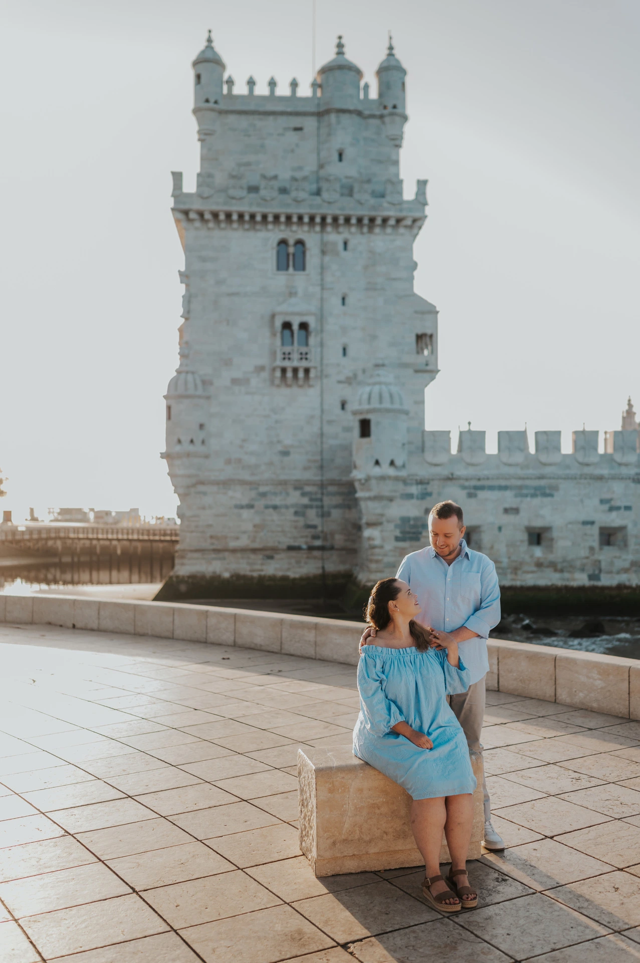 The woman seated on a stone block with her partner close behind her and Belém Tower in the background during their engagement photoshoot in Lisbon.