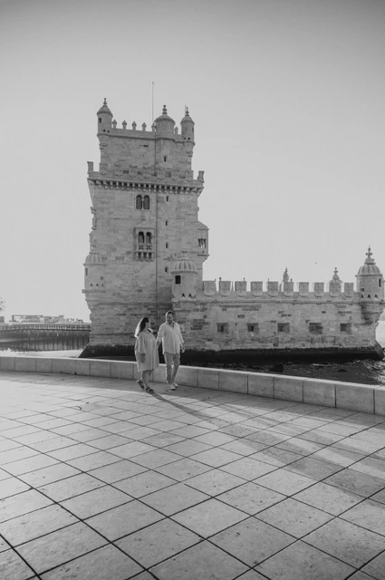 Walking together beside Belém Tower in black and white | Lisbon engagement photoshoot Black-and-white photograph: the couple walking across the waterfront paving with Belém Tower behind them during their engagement photoshoot in Lisbon.