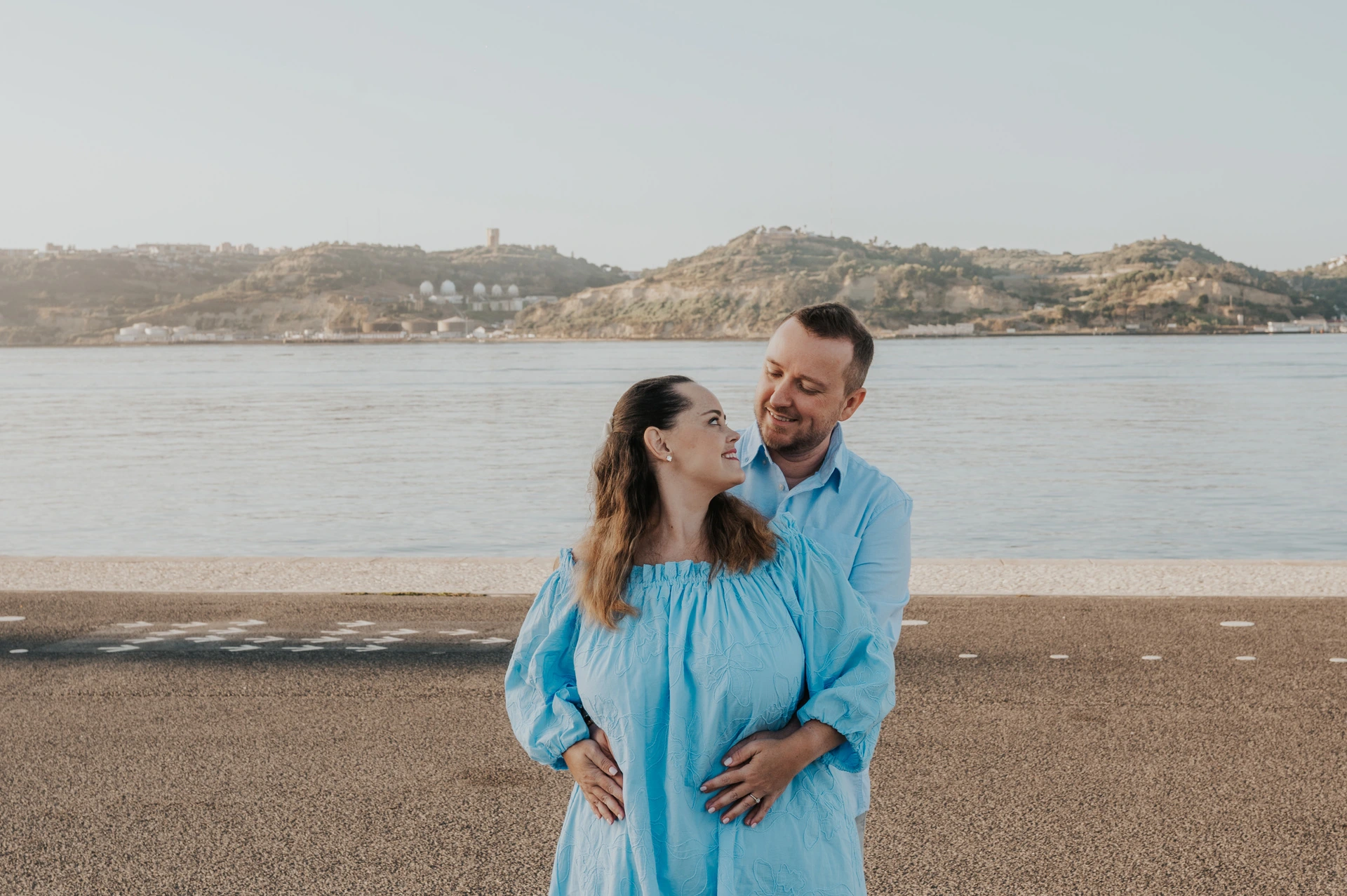 The man embracing his partner from behind beside the river during their engagement photoshoot in Lisbon.