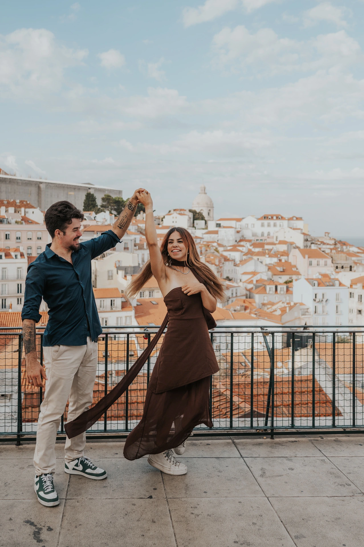 The woman’s brown dress moving as she twirls beside her partner at Portas do Sol during their engagement photoshoot in Lisbon.