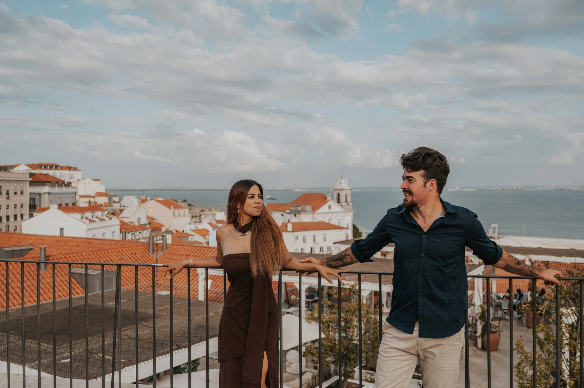 The couple standing apart with their hands on the railing above the rooftops at Portas do Sol during their engagement photoshoot in Lisbon.