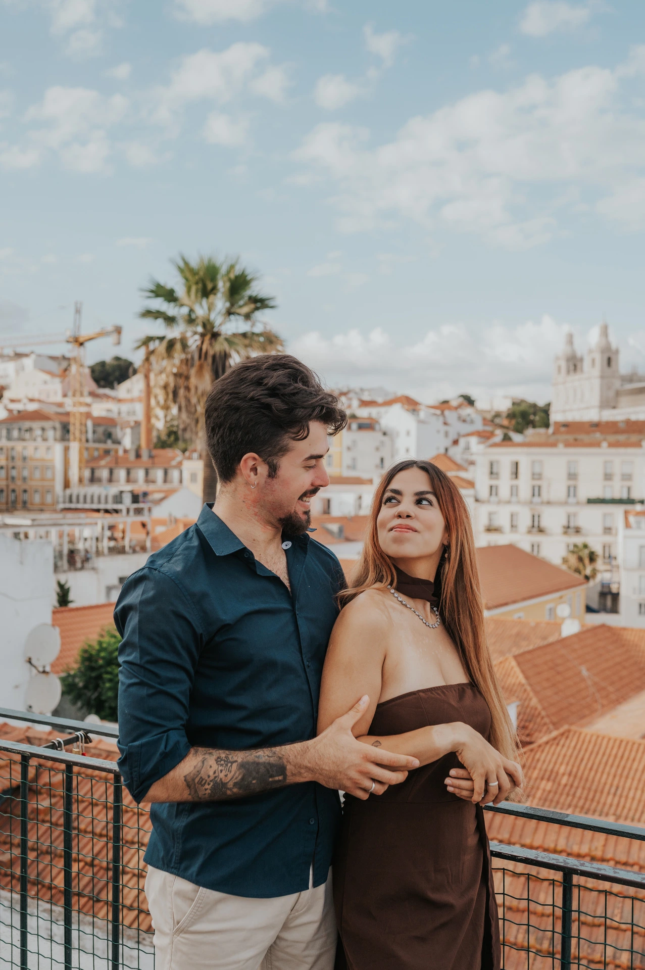 The woman looking back at her partner with a palm tree and rooftops behind them at Portas do Sol during their engagement photoshoot in Lisbon.