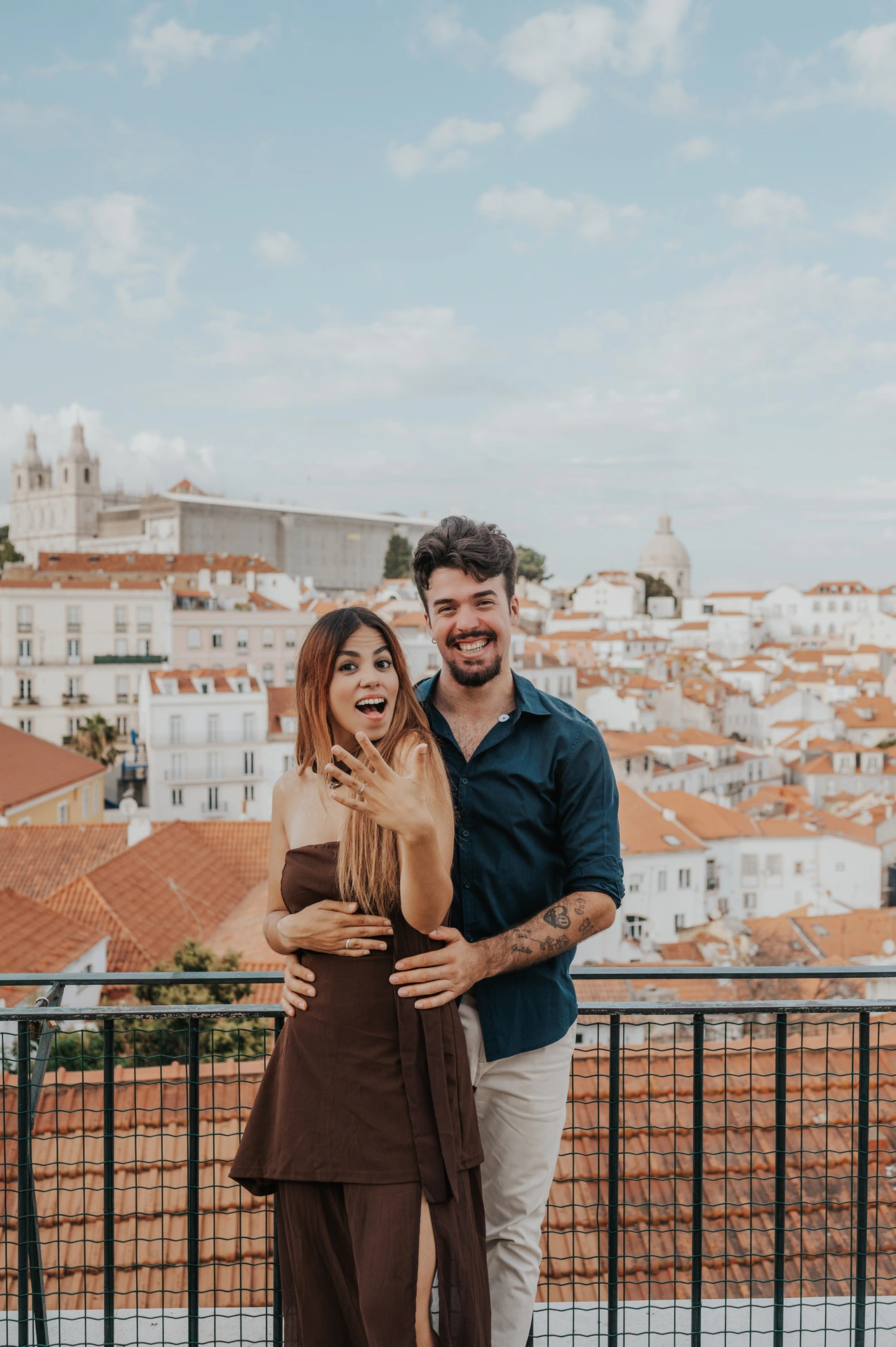 A full-length portrait of the smiling couple showing her engagement ring at Portas do Sol during their engagement photoshoot in Lisbon.