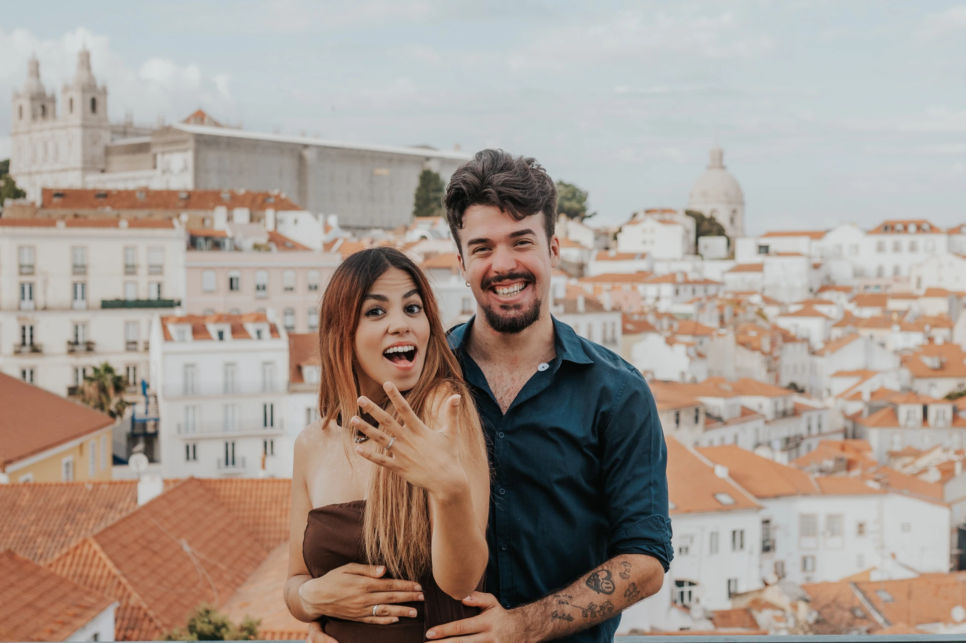Celebrating their engagement above the rooftops | Lisbon engagement photoshoot The couple smiling towards the camera as she holds up her engagement ring at Portas do Sol during their engagement photoshoot in Lisbon.