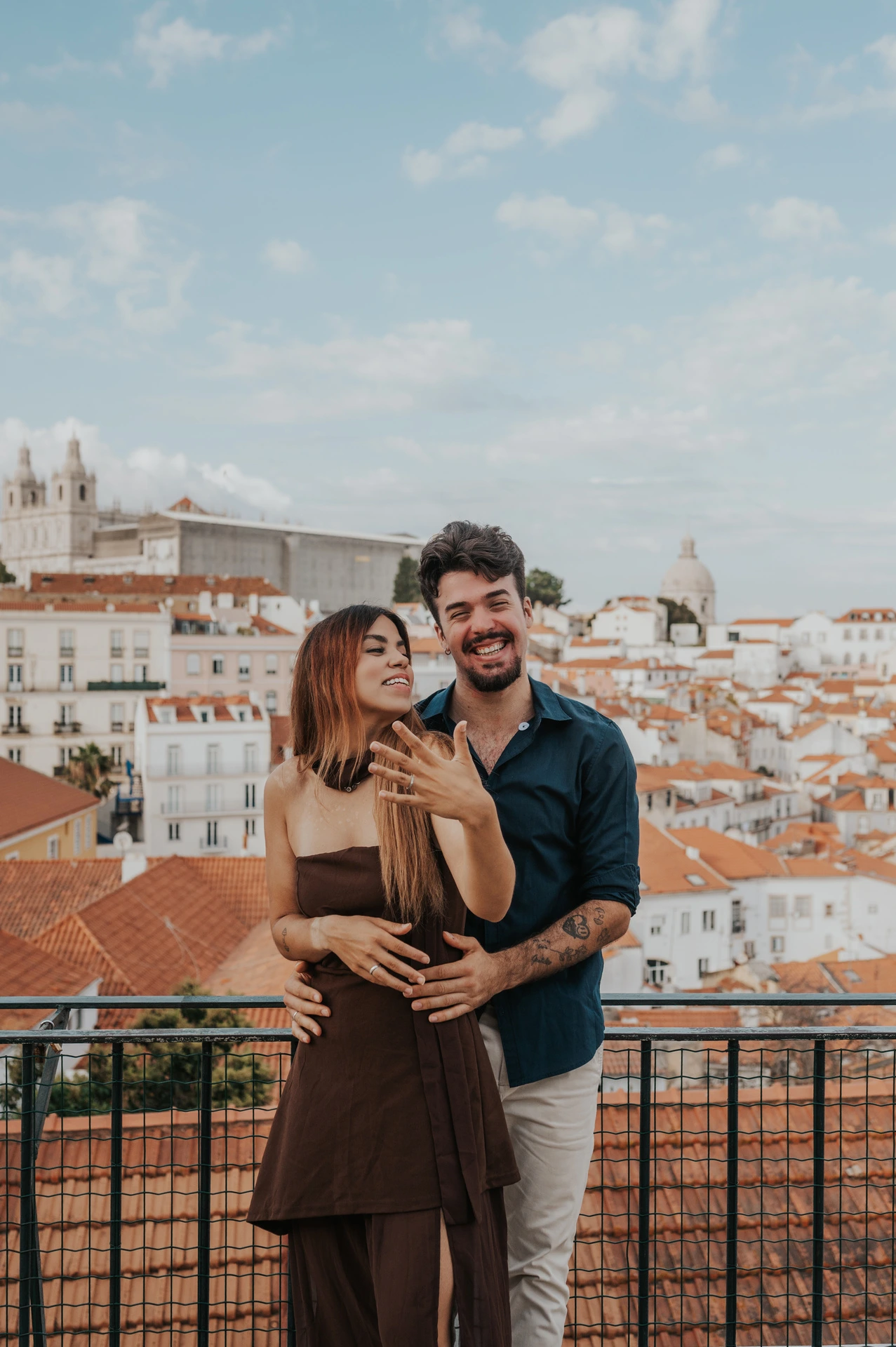 The woman raising her ring hand while her partner smiles behind her at Portas do Sol during their engagement photoshoot in Lisbon.