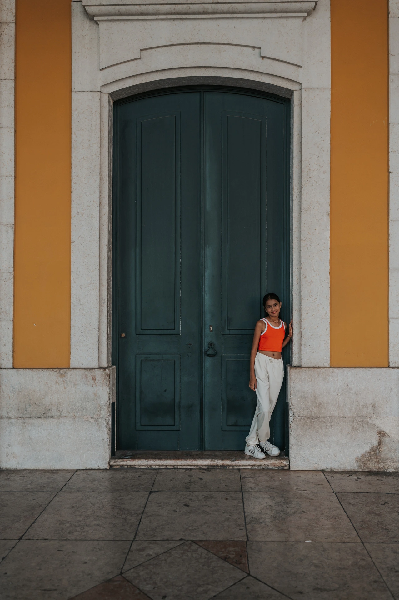 The daughter standing in front of a tall green door framed by the square’s yellow walls during their family and maternity photoshoot in Lisbon.