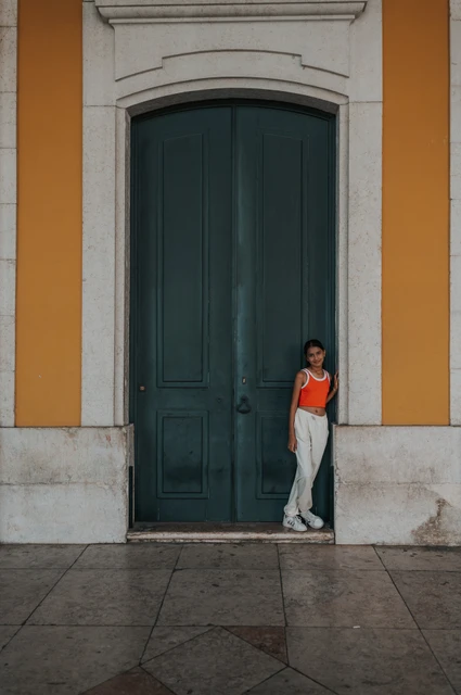The daughter standing in front of a tall green door framed by the square’s yellow walls during their family and maternity photoshoot in Lisbon.