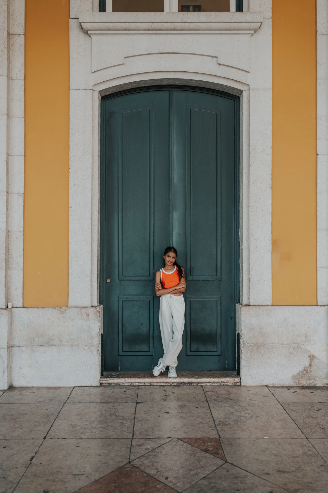 The daughter standing in front of a tall green door framed by the square’s yellow walls during their family and maternity photoshoot in Lisbon.